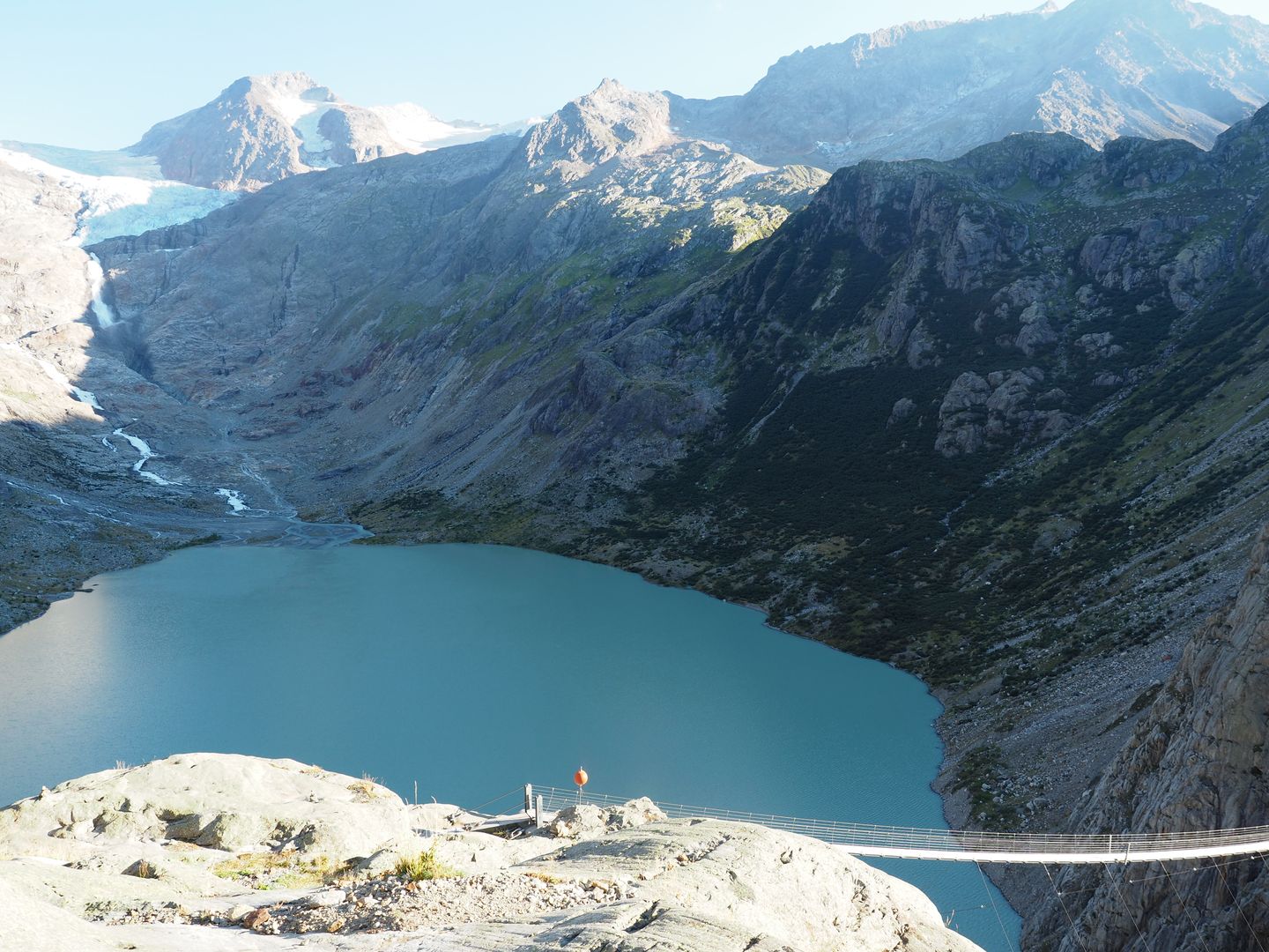 Triftsee mit Gletscher im Hintergrund und Hängebrücke im Vordergrund.