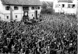 Schwarzweiss Bild, viele Menschen sind auf einem Platz und heben die Hand zum Abstimmen.